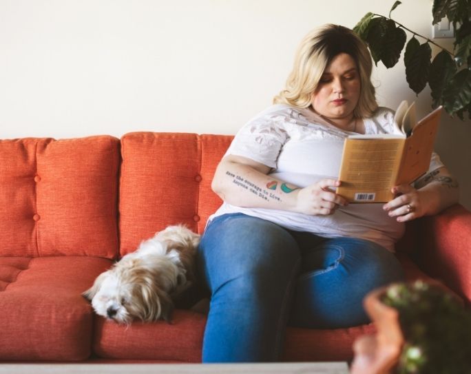 For Pet Sitters from Pet Sitters International A pet sitter with blonde hair wearing a white shirt is sitting on an orange couch reading a book with a small white dog beside her.