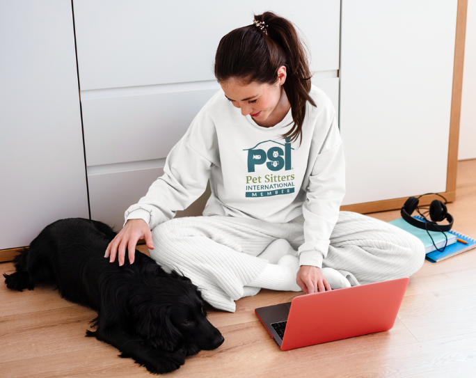 Pet Sitters International Member Benefits Adult woman with dark hair wearing a PSI member sweatshirt pets a dog by her side and looks on laptop