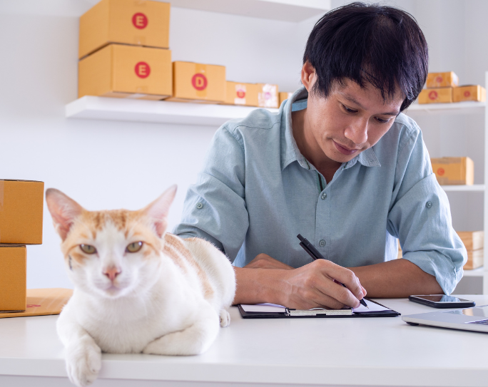 A man in an office with several boxes writes on a clipboard while a cat sits nearby on the desk.